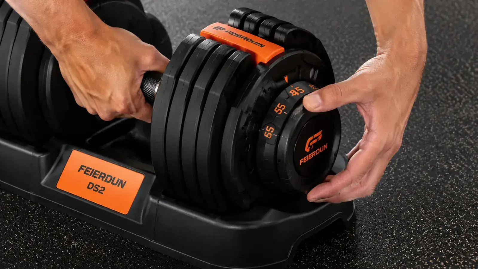 Hands turning the selector dial on a pair of FEIERDUN DS2 adjustable dumbbells set to 45 lbs on a rubber mat