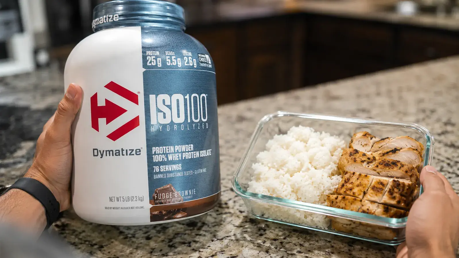 Hand holding a Dymatize ISO100 protein tub next to a meal prep container of grilled chicken and rice