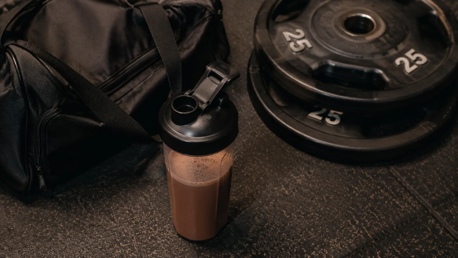 Shaker bottle filled with chocolate protein shake sitting next to a gym bag and weight plates on a home gym floor