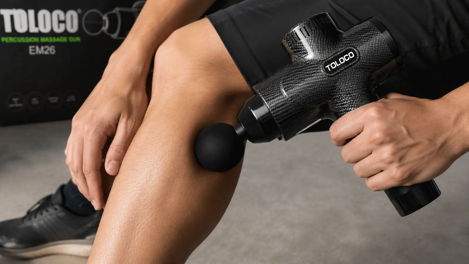 Hand holding the TOLOCO massage gun with the round ball attachment pressed lightly against a calf muscle